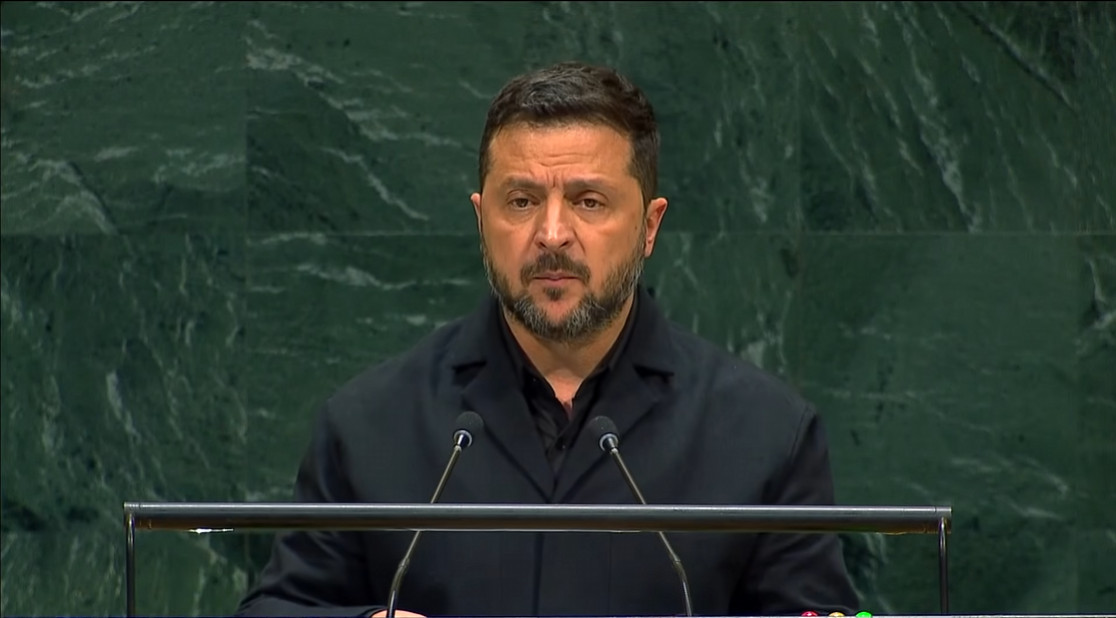 man in a black jacket delivers a speech at a podium with two microphones against a green marble wall